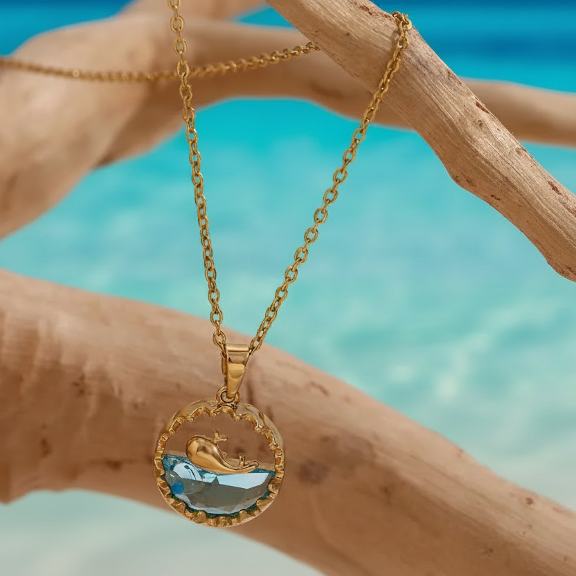 Collier doré en acier inoxydable à fine chaîne maille forçat avec pendentif rond représentant une baleine sur océan en acrylique bleu.