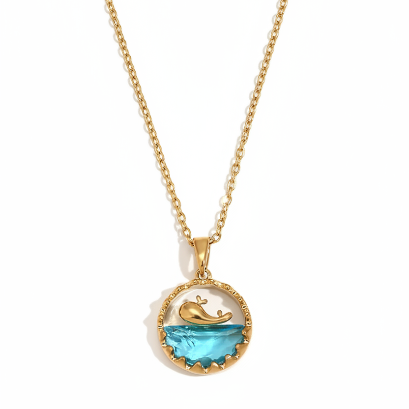 Collier en acier inoxydable doré à fine chaîne maille forçat avec pendentif rond représentant une baleine sur océan en acrylique bleu.