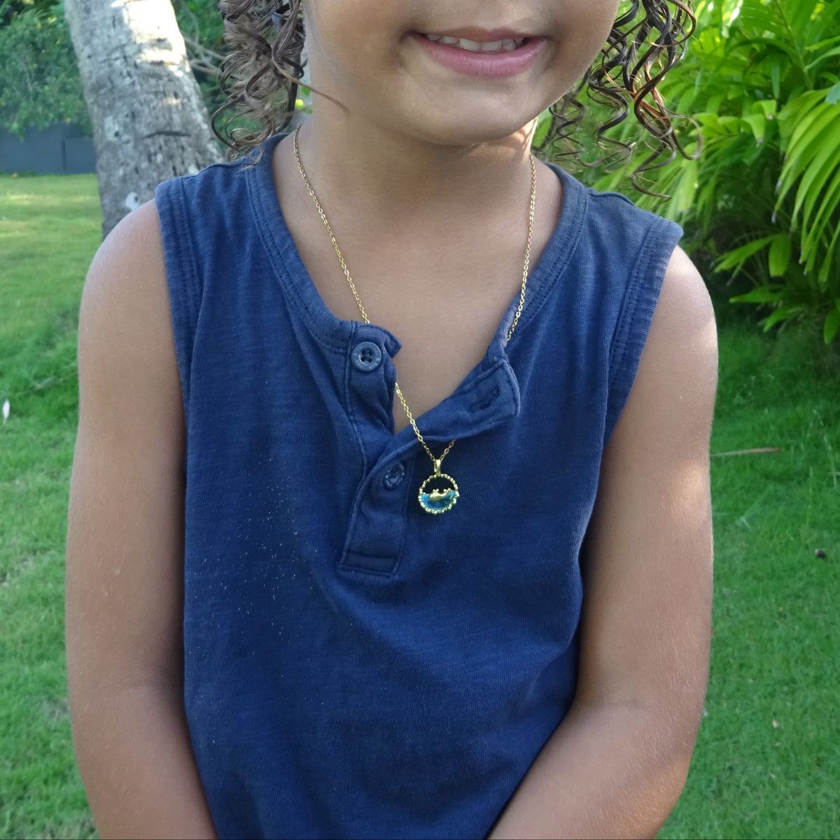 Enfant portant un collier doré en acier inoxydable à fine chaîne maille forçat avec pendentif rond représentant une baleine sur océan en acrylique bleu.